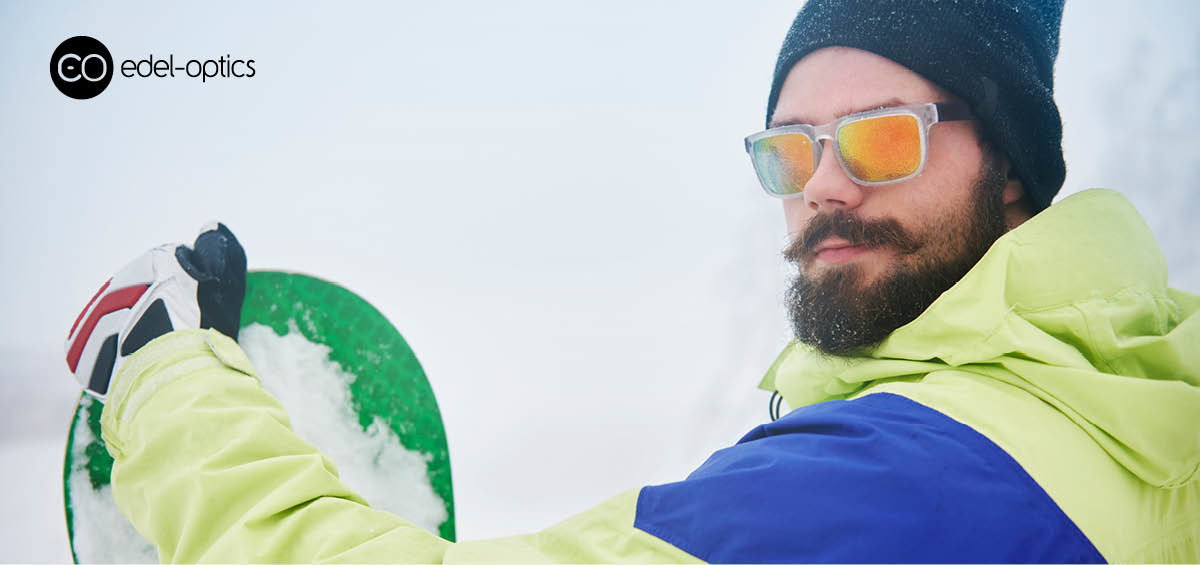 Ein bärtiger Mann mit Ski-Sonnenbrille steht im Schnee und hält ein Snowboard fest.