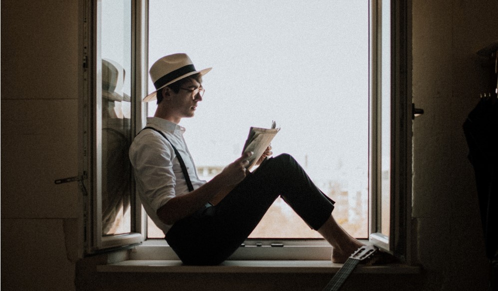 Ein Mann mit Hut und Brille sitzt auf dem Fensterbrett und liest ein Buch.